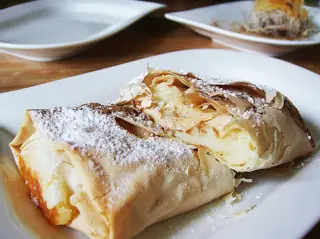 Flaky bougatsa dusted with sugar on a white plate, with another portion and plain tableware behind.