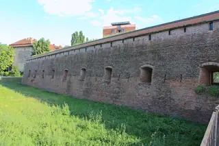 A long brick defensive wall and recessed openings above a grassy foreground, with small structures visible behind.