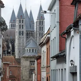 Five stone cathedral towers rise behind a narrow street and nearby red-and-white house façades under gray clouds.