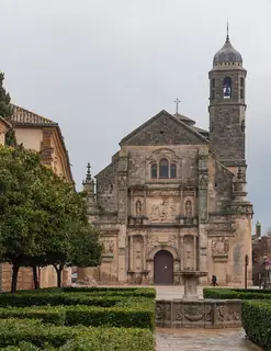 Carved stone chapel facade and bell tower beyond clipped hedges, an orange tree and a tiered stone fountain.