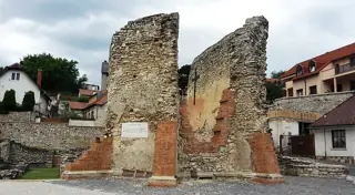 Low stone and brick church ruins, a plain cross and neighboring houses beneath a gray sky.