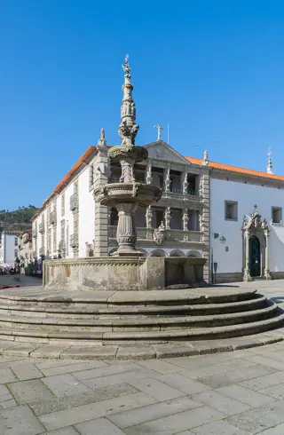 A tiered stone fountain with carved figures in front of an ornate arcaded facade under blue sky.
