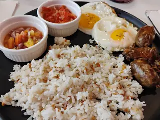 Small browned sausages, fried eggs and rice fill a black plate beside two white bowls of accompaniments.
