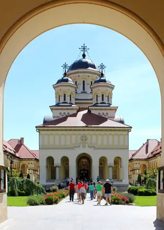 An arch frames a cream-colored cathedral with dark domes, flowerbeds and visitors on the central path.
