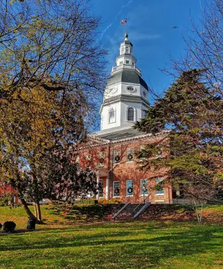 The white tiered dome of Maryland State House rises above a red-brick facade and a tree-lined lawn.