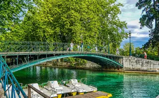 A green metal footbridge over turquoise canal water, with trees and small boats beside the bank.