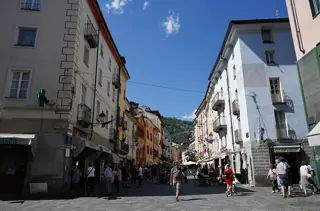 Pedestrians cross a broad paved shopping street between pale façades, with green hills visible beyond the rooftops.