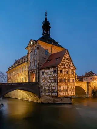 A brightly lit historic building with a tower and half-timbered riverfront section stands beside an arched bridge.
