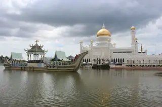 White mosque with a golden dome and slender minaret beside a lagoon, with an ornate roofed ceremonial barge in front under gray clouds.