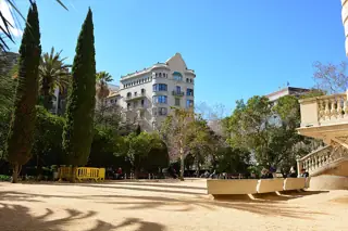 Cypress and palm trees surround an open garden with benches and a stone staircase, backed by city buildings.