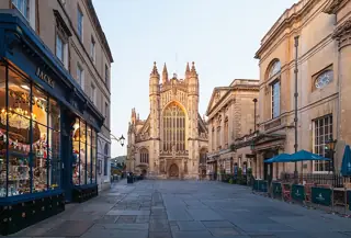 Bath Abbey’s illuminated west front at the end of a paved lane, between shopfronts and the Roman Baths buildings.