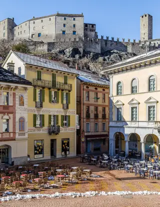 Pastel building fronts and outdoor tables fill a square below castle walls on a rocky rise.