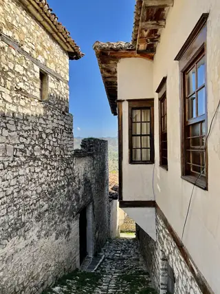 Steep empty cobbled passage between a rough stone wall and a projecting white house with wooden windows.