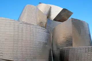 Overlapping silver-grey metal forms rise against a clear blue sky.