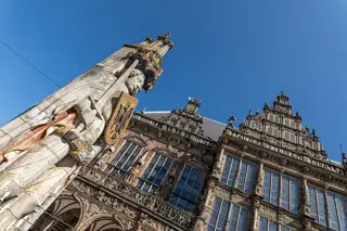 A low-angle photograph looks past the stone Roland statue toward the carved town hall and blue sky.