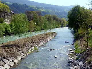 Two pale blue-green rivers meet beside stony banks and dense trees, with wooded hills beyond.