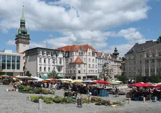 Colourful market awnings and plant trays around a stone fountain, with the Old Town Hall tower behind.