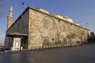 A massive pale-stone mosque wall with pointed window arches, a minaret and domes seen from the public street in Bursa.