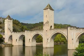 Pale stone towers and pointed arches of Pont Valentré reflected in the Lot beneath a cloudy sky.