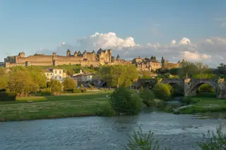 The river Aude and a stone bridge below a sunlit hilltop city of crenellated walls and pointed towers.
