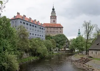 A painted castle and round tower rise above trees and a curving river, with cobbled banks below.