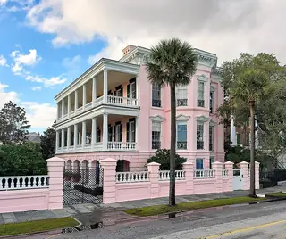 Pink three-storey Charleston house with broad porches, iron railings and palms, with scaffolding visible at the right edge.