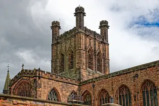 A square stone tower with corner turrets rises above Gothic windows beneath a cloudy sky.