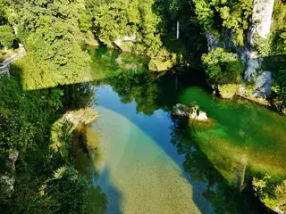 Clear green river water reflects trees between pale rock banks, with a small plain terrace at the left and dark steps at the rear.