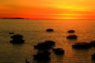 Dark boat silhouettes float on rippling water beneath an orange sunset and distant low hills.