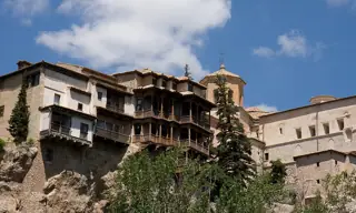 Dark wooden balconies project from pale houses above a rocky slope, framed by trees and blue sky.