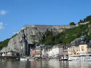 The Meuse reflects Dinant’s riverside buildings, with a dark onion-domed church beneath a cliff-top stone citadel.