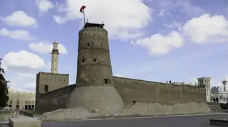 A broad sandy-stone fort wall and round defensive tower beneath a blue sky, with mosque minarets and smaller structures visible behind.