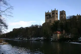 Snowy branches and a shaded river frame Durham Cathedral and riverside buildings.