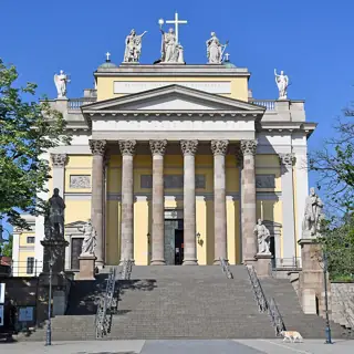 Six tall columns front a yellow basilica above stone steps, with figures along the stairway and roof against blue sky.