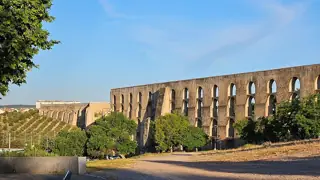 Rows of stone aqueduct arches cross a shallow valley above trees and dry open ground beneath a blue sky.