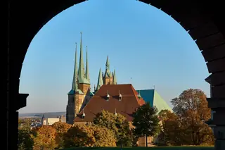 Green church spires and steep red roofs rise beyond autumn trees, framed by a dark stone arch.
