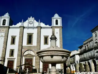 A broad marble fountain bowl stands before a white church facade and arcaded buildings under a blue sky.
