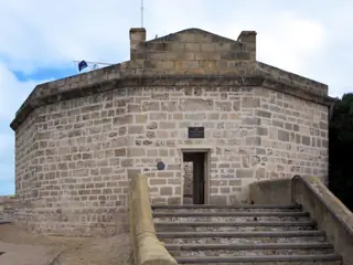 A low many-sided limestone building faces a broad flight of stone steps, with an open doorway beneath a small sign.