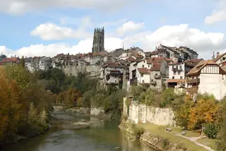 River curving below autumn trees, stone cliffs and closely packed houses beneath a Gothic tower.