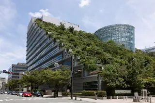 A stepped modern building is covered in dense greenery beside a glass atrium, with a street crossing and vehicles below.