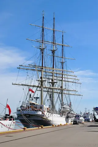 A white-hulled sailing ship with three tall masts and dense rigging is moored beside a paved quay beneath blue sky.