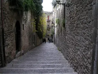 A steep cobbled staircase descends between rough stone walls, greenery and iron lamps, with a few distant pedestrians.