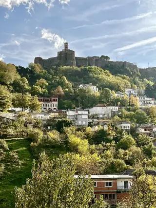 A long stone fortress and clock tower crown a green hillside dotted with white houses.