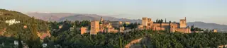 A wide evening panorama of warm stone towers and palaces above a wooded slope, with mountains behind.