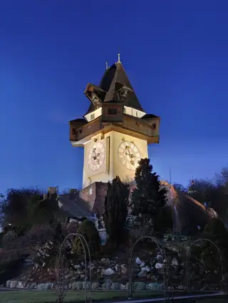 A pale clock tower with a dark wooden balcony rises above trees against a deep blue evening sky.