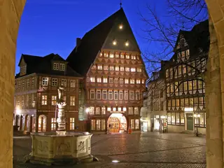 Illuminated half-timbered guildhall behind a carved stone fountain, framed by a stone arcade.