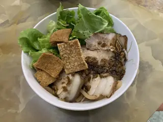 Sliced pork, leafy greens and crisp golden pieces cover a white bowl on a mottled tabletop.