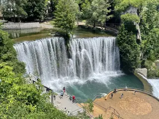 A broad curtain of water falls into a green pool beside trees, stone banks and a lower viewing platform.