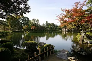 Red and green trees surround a reflective pond, with a stone lantern at right and a small pavilion across the water.