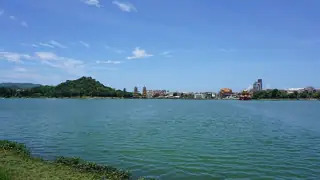 Blue-green lake water stretches toward a wooded hill, distant pagodas and low city buildings beneath a broad blue sky.
